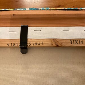 May include: Close-up of the back of a wooden canvas frame with white fabric stretched across it. The frame has a black metal hanging clip and the numbers "9121", "51801", and "11X14" printed on it.