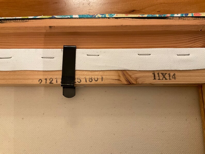 May include: Close-up of the back of a wooden canvas frame with white fabric stretched across it. The frame has a black metal hanging clip and the numbers "9121", "51801", and "11X14" printed on it.