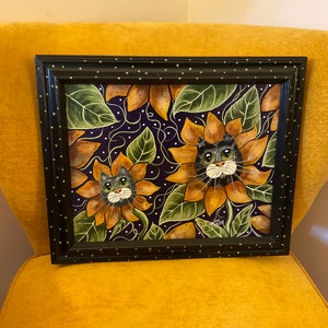 May include: A framed painting of two black cats peeking out of sunflowers. The sunflowers are yellow and brown with green leaves. The background is a dark purple with white dots.