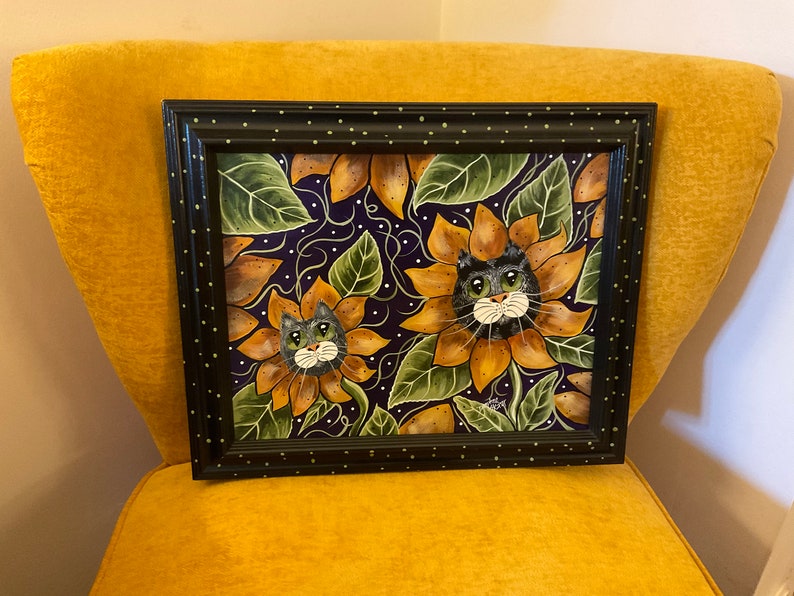 May include: A framed painting of two black cats peeking out of sunflowers. The sunflowers are yellow and brown with green leaves. The background is a dark purple with white dots.