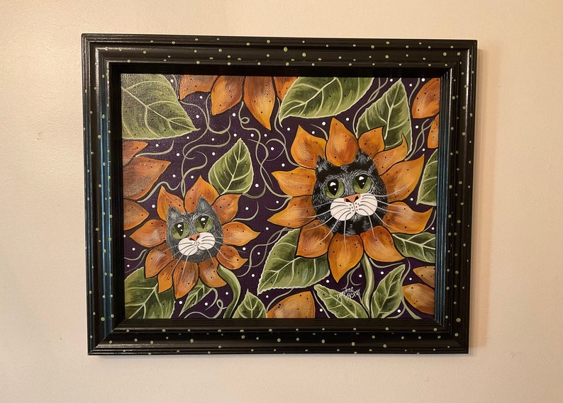 May include: A framed painting of two black cats with white faces peeking out of sunflowers. The sunflowers are orange and yellow with green leaves. The background is a dark purple with white dots.