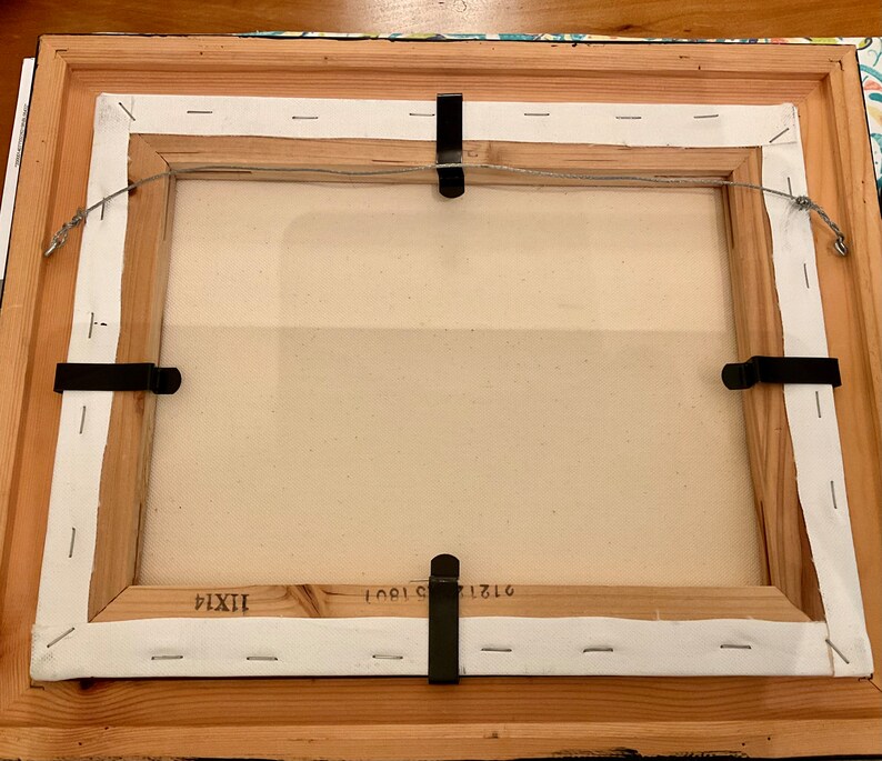 May include: The back of a framed canvas painting with a white canvas and a wooden frame. The frame has a white paper backing with staples and a wire attached to the top for hanging. The canvas is labeled with the numbers "1710", "1901", and "IXXI".