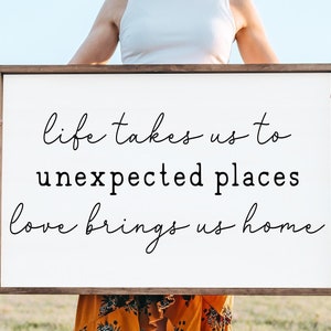 May include: A white wooden sign with the text "life takes us to unexpected places love brings us home" written in black cursive font.