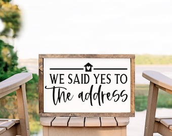 May include: A wooden sign with a white background and black text that reads "We said yes to the address". The sign is sitting on a small wooden stool between two wooden rocking chairs. The chairs are facing the water.