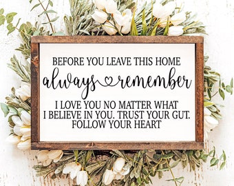 May include: A wooden sign with a white background and black text that reads "Before you leave this home always remember I love you no matter what I believe in you. Trust your gut. Follow your heart." The sign is surrounded by white flowers and green leaves.