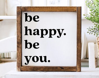 May include: A wooden sign with the text "be happy. be you." in black lettering on a white background.
