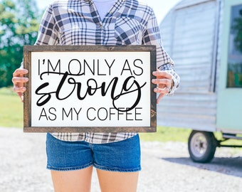 May include: A wooden sign with the text "I'm only as strong as my coffee" in black script font. The sign is held by a person wearing a plaid shirt and denim shorts.