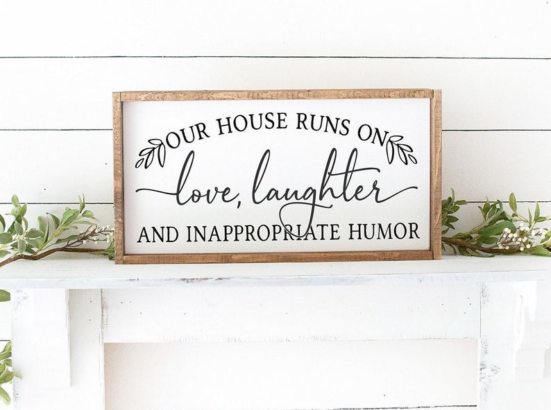 Pu&ograve; includere: Un cartello di legno con il testo "Our house runs on love, laughter and inappropriate humor" in lettere nere. Il cartello &egrave; su uno scaffale bianco con piante verdi su entrambi i lati.