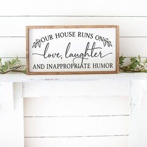 Pu&ograve; includere: Un cartello di legno con il testo "Our house runs on love, laughter and inappropriate humor" in corsivo nero. Il cartello &egrave; su un camino bianco con rami verdi e fogliame su entrambi i lati.