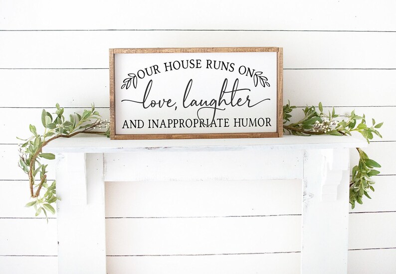 Pu&ograve; includere: Un cartello di legno con il testo "Our house runs on love, laughter and inappropriate humor" in corsivo nero. Il cartello &egrave; su un camino bianco con rami verdi e fogliame su entrambi i lati.