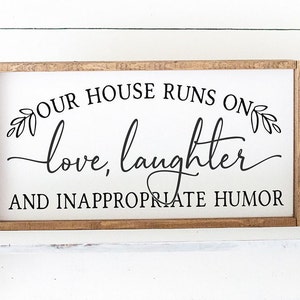 Pu&ograve; includere: Un cartello in legno bianco con cornice marrone recita "La nostra casa funziona con amore, risate e umorismo inappropriato". Il cartello &egrave; decorato con piccoli disegni di foglie.