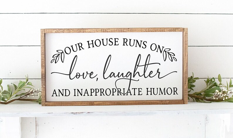 Pu&ograve; includere: Un cartello in legno bianco con cornice marrone recita "La nostra casa funziona con amore, risate e umorismo inappropriato". Il cartello &egrave; decorato con piccoli disegni di foglie.