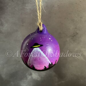 Pode incluir: Ornamento de cerâmica pintado à mão em roxo e rosa com uma montanha branca e um disco voador no céu. O ornamento tem uma corda de juta para pendurar.