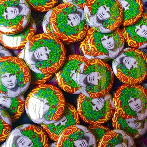 Puede incluir: Una colección de botones redondos y coloridos que presentan una imagen psicodélica de una persona con una corona floral. Los botones tienen un fondo naranja y amarillo en espiral con flores verdes y azules.