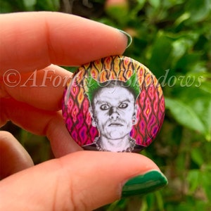 Puede incluir: Un botón redondo con un diseño psicodélico colorido que presenta un retrato en blanco y negro de una persona con cabello verde. El fondo es un patrón de remolino de rojo, naranja, amarillo y rosa.