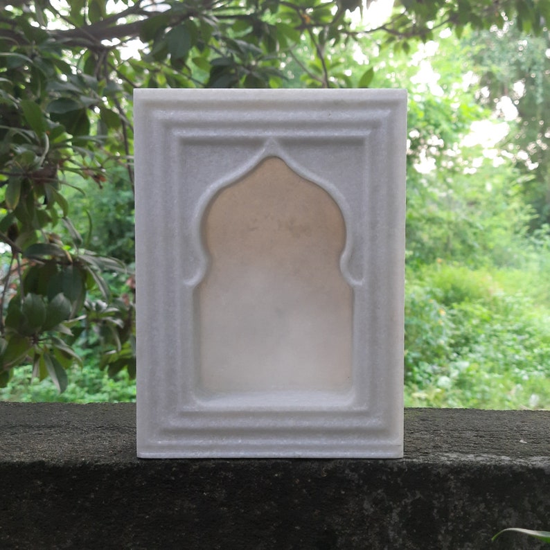 May include: A white marble decorative frame with an arched interior. The frame has a rectangular shape with a detailed border. The background is a blurred view of green foliage.