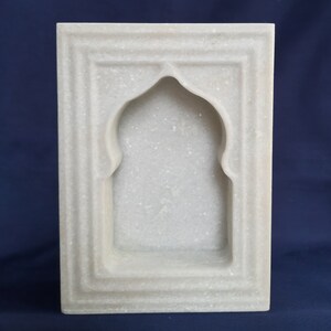 May include: A rectangular, white marble wall niche with an arched top. The niche is framed by a series of recessed borders, creating a layered effect. The marble has a subtle, speckled texture.