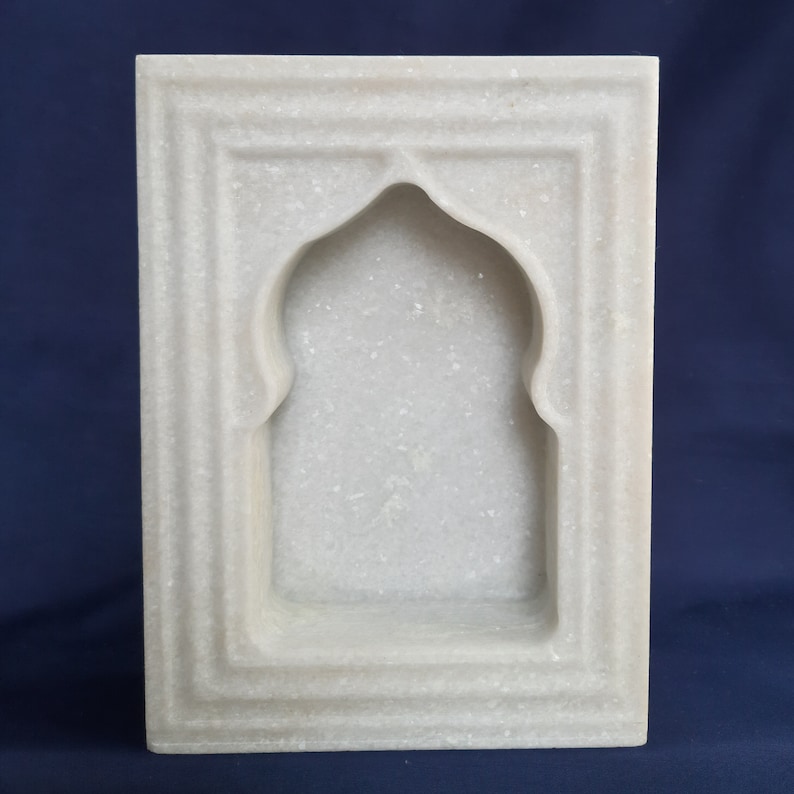 May include: A rectangular, white marble wall niche with an arched top. The niche is framed by a series of recessed borders, creating a layered effect. The marble has a subtle, speckled texture.