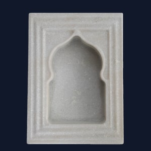 May include: A white marble wall niche with an arched top and rectangular frame. The niche has a recessed interior and is designed with a layered, textured border. The background is a dark blue.