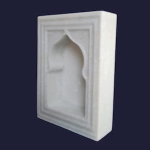 May include: A white marble decorative wall hanging with an arched niche design. The rectangular piece features a recessed archway and a framed border, showcasing a minimalist aesthetic. The smooth, polished surface reflects light, highlighting the natural veining of the stone.
