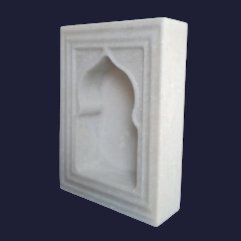 May include: A white marble decorative wall hanging with an arched niche design. The rectangular piece features a recessed archway and a framed border, showcasing a minimalist aesthetic. The smooth, polished surface reflects light, highlighting the natural veining of the stone.