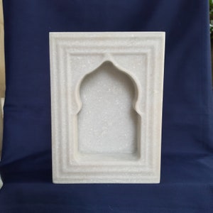 May include: A rectangular, white marble wall niche with an arched top. The niche is framed by a series of recessed borders, creating a layered effect. The overall design is simple and elegant, suitable for displaying small objects.