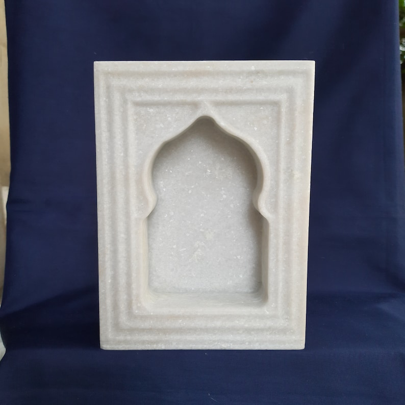 May include: A rectangular, white marble wall niche with an arched top. The niche is framed by a series of recessed borders, creating a layered effect. The overall design is simple and elegant, suitable for displaying small objects.
