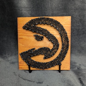 Puede incluir: Un letrero de madera con un diseño de flecha de arte de hilo negro. La flecha está hecha con hilo negro envuelto alrededor de pequeños clavos en una tabla de madera marrón claro. El letrero se muestra en un soporte de metal negro.