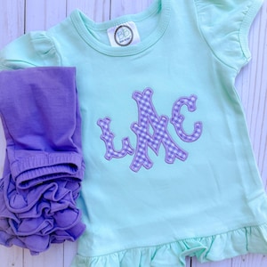 May include: A light blue baby t-shirt with a ruffled hem and a purple gingham monogram "LMC" on the front. The shirt is paired with purple ruffle leggings.