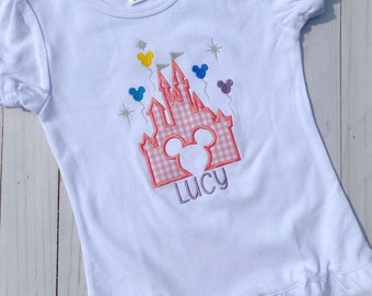 Puede incluir: Camiseta blanca con un diseño de castillo de vichy rosa y blanco, orejas de Mickey Mouse y globos. El nombre "Lucy" está bordado debajo del castillo.