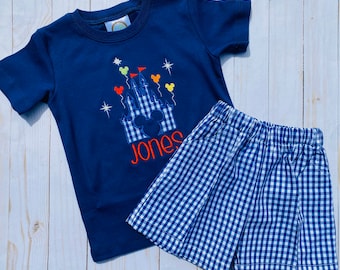 May include: A navy blue t-shirt featuring an embroidered image of a castle with Mickey Mouse ears and the name "Jones" underneath. The shirt is paired with blue and white checked shorts.