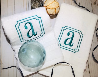 May include: White bath towels with a teal embroidered letter "A" inside a decorative frame. The towels are folded and arranged with decorative glass and wooden spheres, and a dark ribbon on a light wood surface.