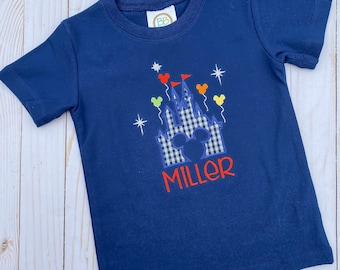 Puede incluir: Una camiseta azul marino con un diseño de castillo a cuadros blancos y azules, orejas de Mickey Mouse y globos de colores. El nombre "MILLER" está bordado en rojo debajo del castillo.