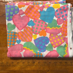 May include: Folded fabric with a white background and a pattern of colorful hearts. The hearts are in various colors, including pink, blue, green, and orange, and feature polka dots, stripes, and checkered designs. The fabric is folded neatly.