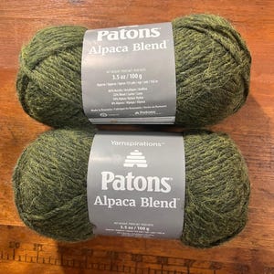 May include: Two skeins of olive green Patons Alpaca Blend yarn, each weighing 3.5 ounces. The labels state a composition of 60% acrylic, 22% wool, 10% nylon, and 8% alpaca, with a length of approximately 155 yards per 100 gram skein.