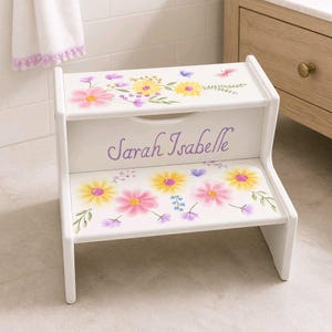May include: A white two-step stool with floral designs in pink, yellow, and purple. The name "Sarah Isabelle" is written in purple script on the front. The stool is designed for children and has a decorative, painted finish.