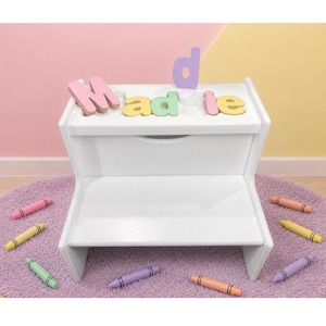 May include: A white two-step stool with a smooth finish. The top step features pastel-colored wooden letters spelling out the name Maddie. Several colorful crayons are scattered around the base of the stool on a circular purple rug.
