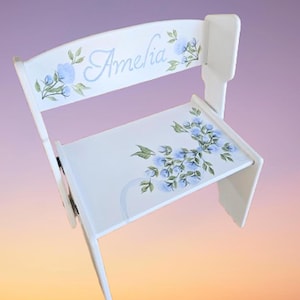 May include: A white wooden child's chair with a backrest and seat. The backrest is adorned with the name "Amelia" in script, along with blue floral accents. The seat also features blue floral designs. The chair is painted white.