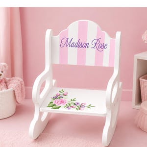K&ouml;nnte beinhalten: Ein wei&szlig;er Schaukelstuhl aus Holz mit einer R&uuml;ckenlehne mit vertikalen hellrosa Streifen und dem in violetter Schreibschrift aufgedruckten Namen Madison Rose. Die Sitzfl&auml;che ist mit einem Blumenmuster aus rosa Rosen und gr&uuml;nen Bl&auml;ttern verziert.