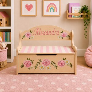 May include: A light-colored wooden toy chest bench with a backrest featuring the name Alexandra in pink script. The piece is decorated with pink floral motifs and has a pink and white striped seat. The front panel displays a floral arrangement.