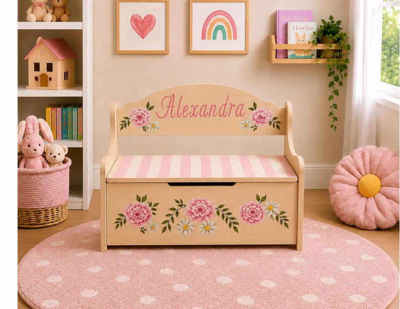 May include: A light-colored wooden toy chest bench with a backrest featuring the name Alexandra in pink script. The piece is decorated with pink floral motifs and has a pink and white striped seat. The front panel displays a floral arrangement.