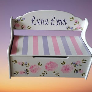 May include: White wooden storage bench with a pink and purple striped seat and floral painted design. The bench is personalized with the name "Luna Lynn".
