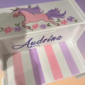 May include: White wooden step stool with a pink and purple unicorn painted on the top. The stool has a purple and pink striped design on the step. The name "Audrina" is painted on the front of the stool.