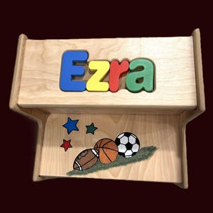 May include: Wooden step stool with the name "Ezra" in blue, yellow, and red letters. The lower section features painted sports balls and stars. The stool is made of light-colored wood.