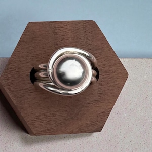 Op de afbeelding: Een zilveren ring met een grote, ronde, gepolijste middensteen. De band van de ring heeft een wervelend ontwerp dat zich om de centrale steen wikkelt. De ring wordt gepresenteerd op een bruine, zeshoekige houten standaard.
