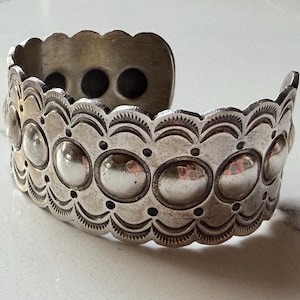 May include: A silver-toned metal cuff bracelet featuring a scalloped edge and a central row of raised, polished circular domes. The surface is decorated with stamped semi-circular patterns and small dots, with three dark circular magnets visible inside.