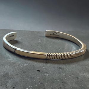 Puede incluir: Una pulsera de tipo brazalete en plata que presenta una sección lisa pulida y un segmento texturizado con líneas verticales ranuradas. El interior de la banda está grabado con el texto MK STERLING, indicando la composición de este metal.