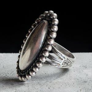 May include: A silver ring featuring a large, smooth, elongated oval centerpiece surrounded by a border of small silver spheres. The band is split into three sections and displays engraved geometric arrow and line patterns on the sides.