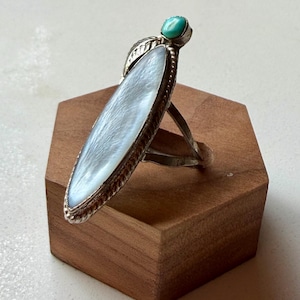 May include: A long, marquise-shaped ring featuring a large, iridescent white mother-of-pearl inlay set in a silver frame with a twisted rope border. A small, round, bright blue turquoise cabochon is positioned at the top of the silver setting.