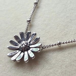 May include: A silver-toned necklace featuring a daisy-shaped pendant with elongated petals and a textured, beaded center. The pendant is attached to a delicate chain that includes small, evenly spaced metal beads along its length.